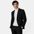 Young man in a black suit with a cream turtleneck sweater, posing with hands in pockets.