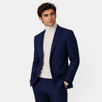 Young man in a navy blue suit with a cream turtleneck sweater, posing with hands in pockets.