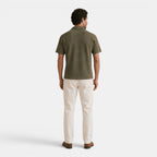 Back view of a man wearing an olive green textured knit polo shirt, cream trousers, and brown loafers against a light gray background.