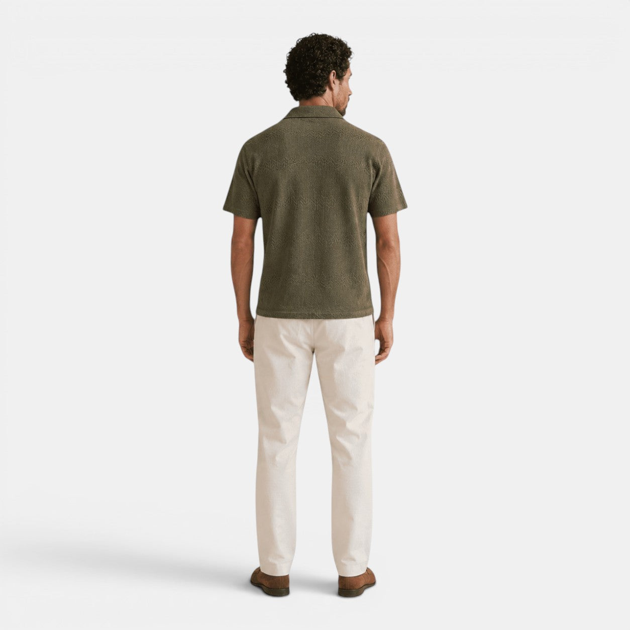 Back view of a man wearing an olive green textured knit polo shirt, cream trousers, and brown loafers against a light gray background.