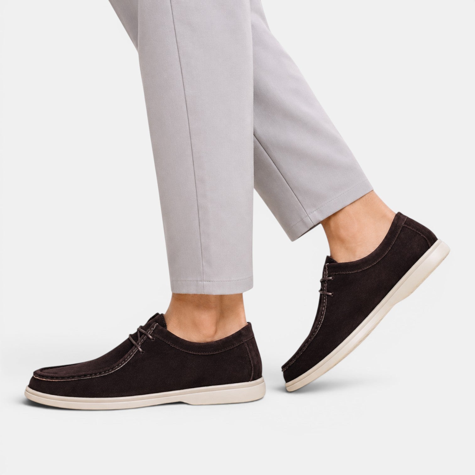 Person wearing dark brown suede lace-up shoes with white soles and light gray trousers, shown in a side walking pose.