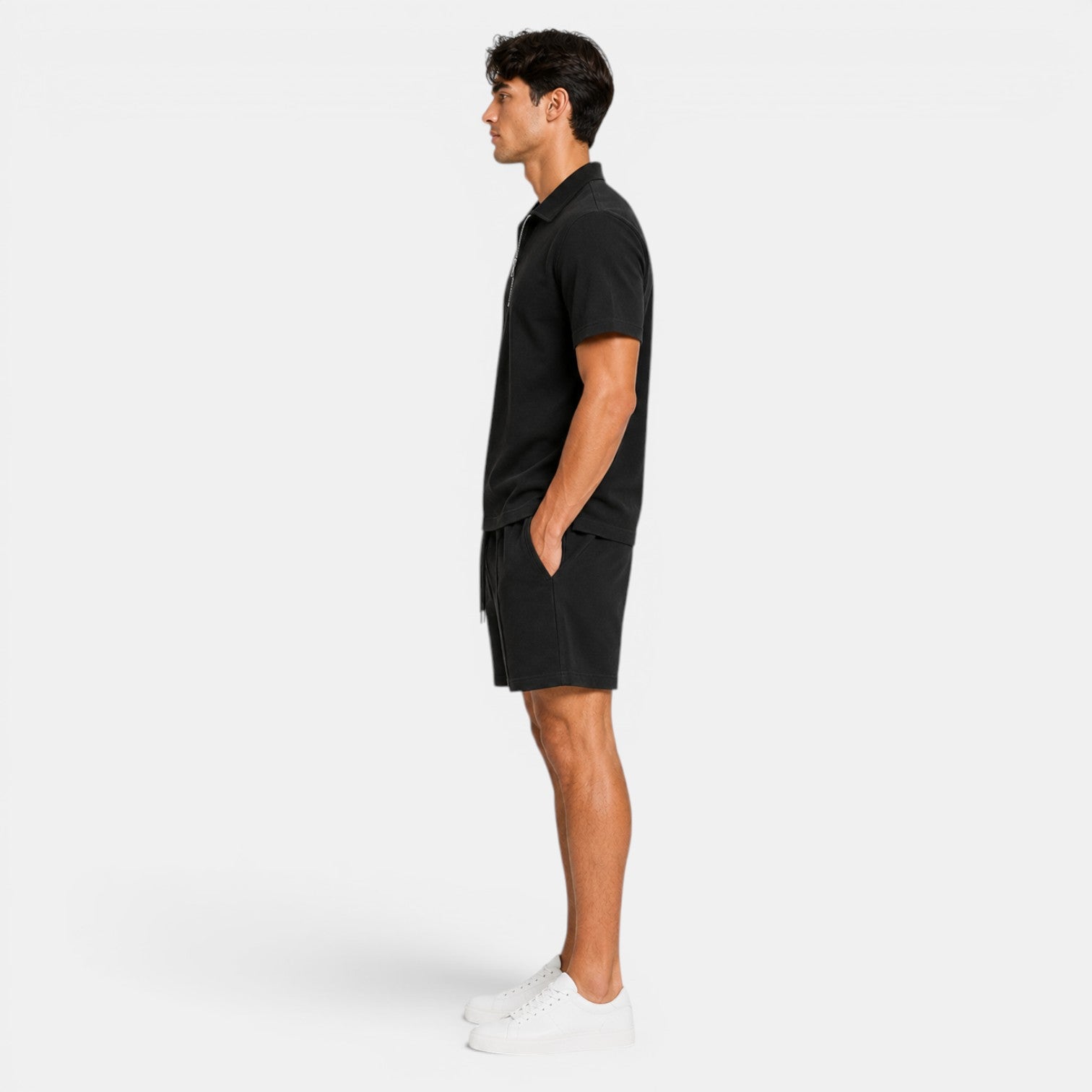 Side view of a man wearing a black zip-collar polo shirt, matching shorts, and white sneakers against a light gray background.