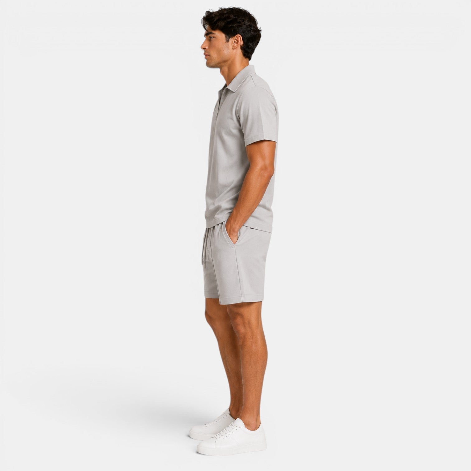 Side view of a man wearing a light gray zip-collar polo shirt, matching shorts, and white sneakers against a light gray background.