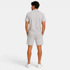Back view of a man wearing a light gray polo shirt, matching shorts, and white sneakers against a light gray background.