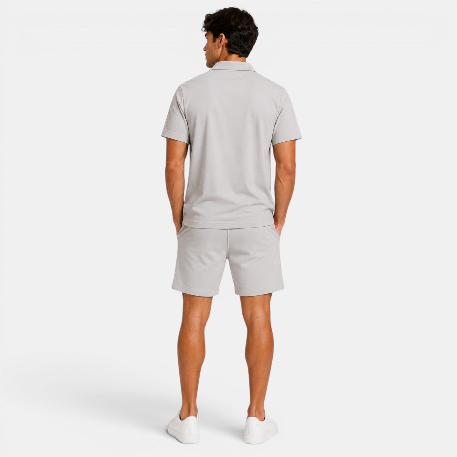 Back view of a man wearing a light gray polo shirt, matching shorts, and white sneakers against a light gray background.