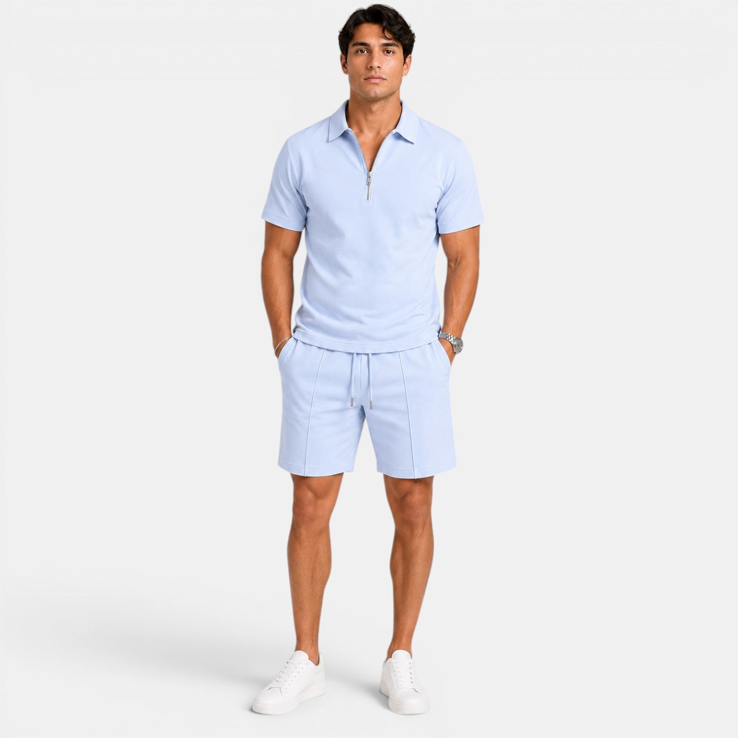 Man wearing a light blue zip-collar polo shirt with matching drawstring shorts and white sneakers, standing against a light gray background.