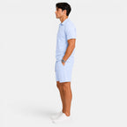 Side view of a man wearing a light blue zip-collar polo shirt, matching shorts, and white sneakers against a light gray background.