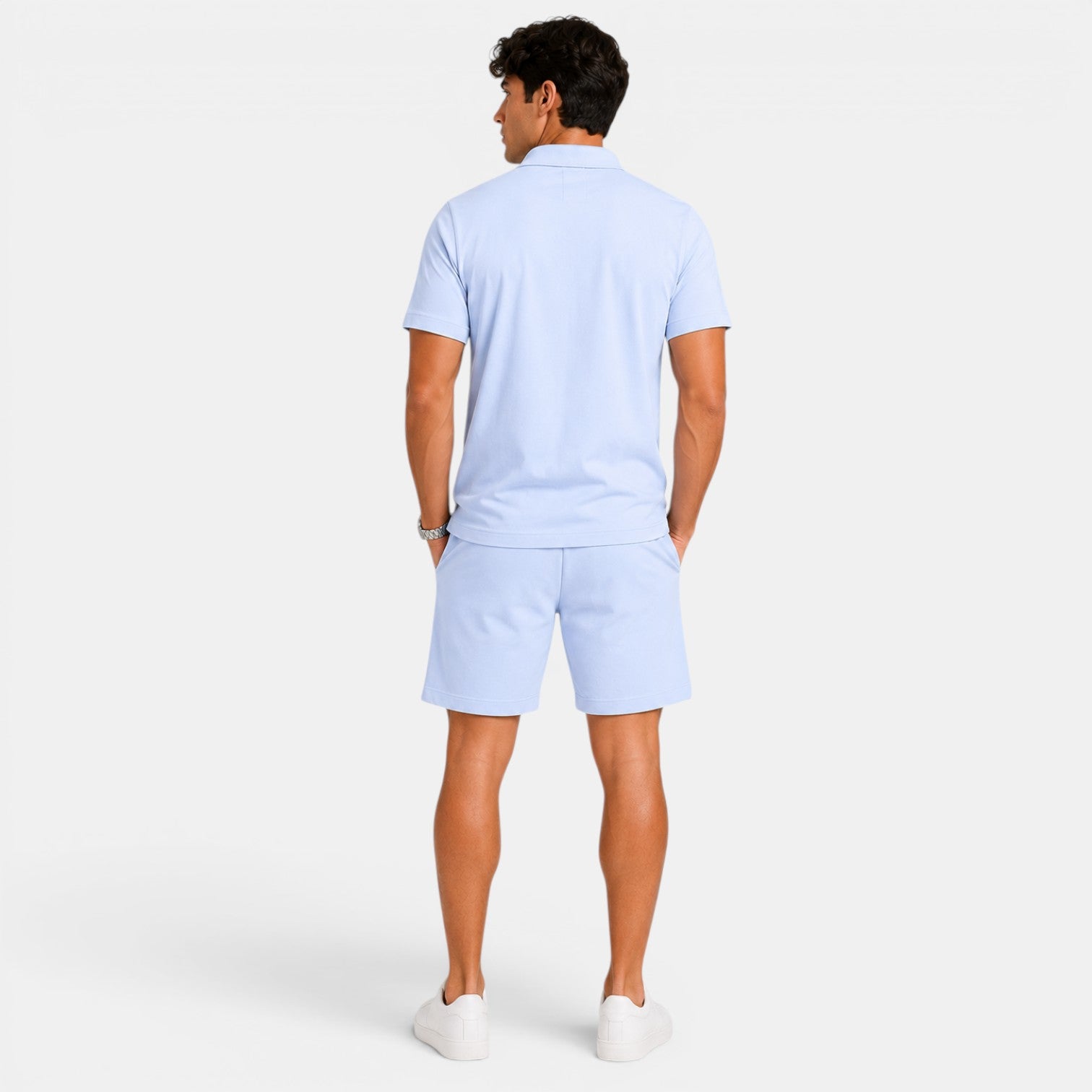Back view of a man wearing a light blue polo shirt, matching shorts, and white sneakers against a light gray background.