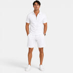 Man wearing a white zip-collar polo shirt with matching drawstring shorts and white sneakers, standing against a light gray background.