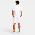 Back view of a man wearing a white polo shirt, matching shorts, and white sneakers against a light gray background.
