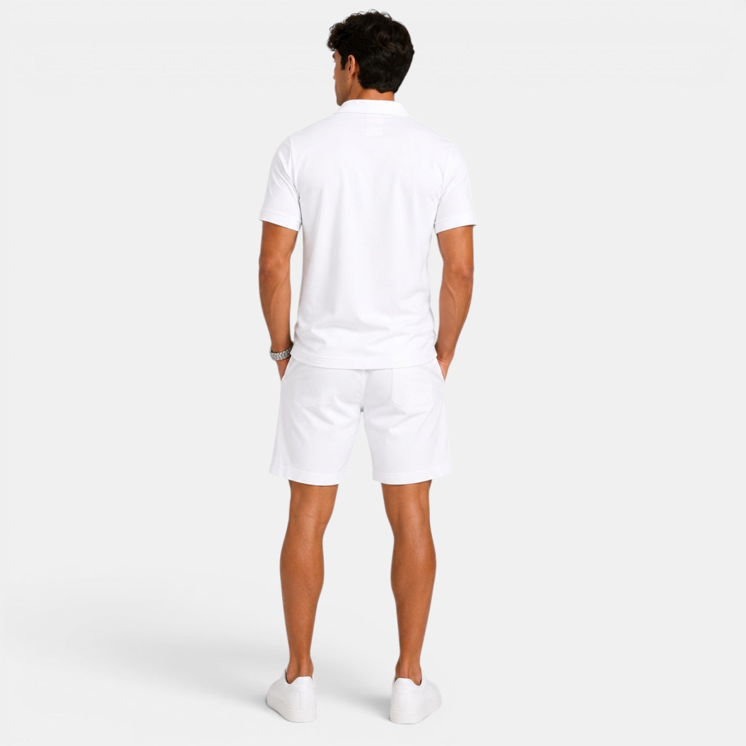 Back view of a man wearing a white polo shirt, matching shorts, and white sneakers against a light gray background.