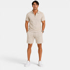 Man wearing a beige zip-collar polo shirt with matching drawstring shorts and white sneakers, standing against a light gray background.