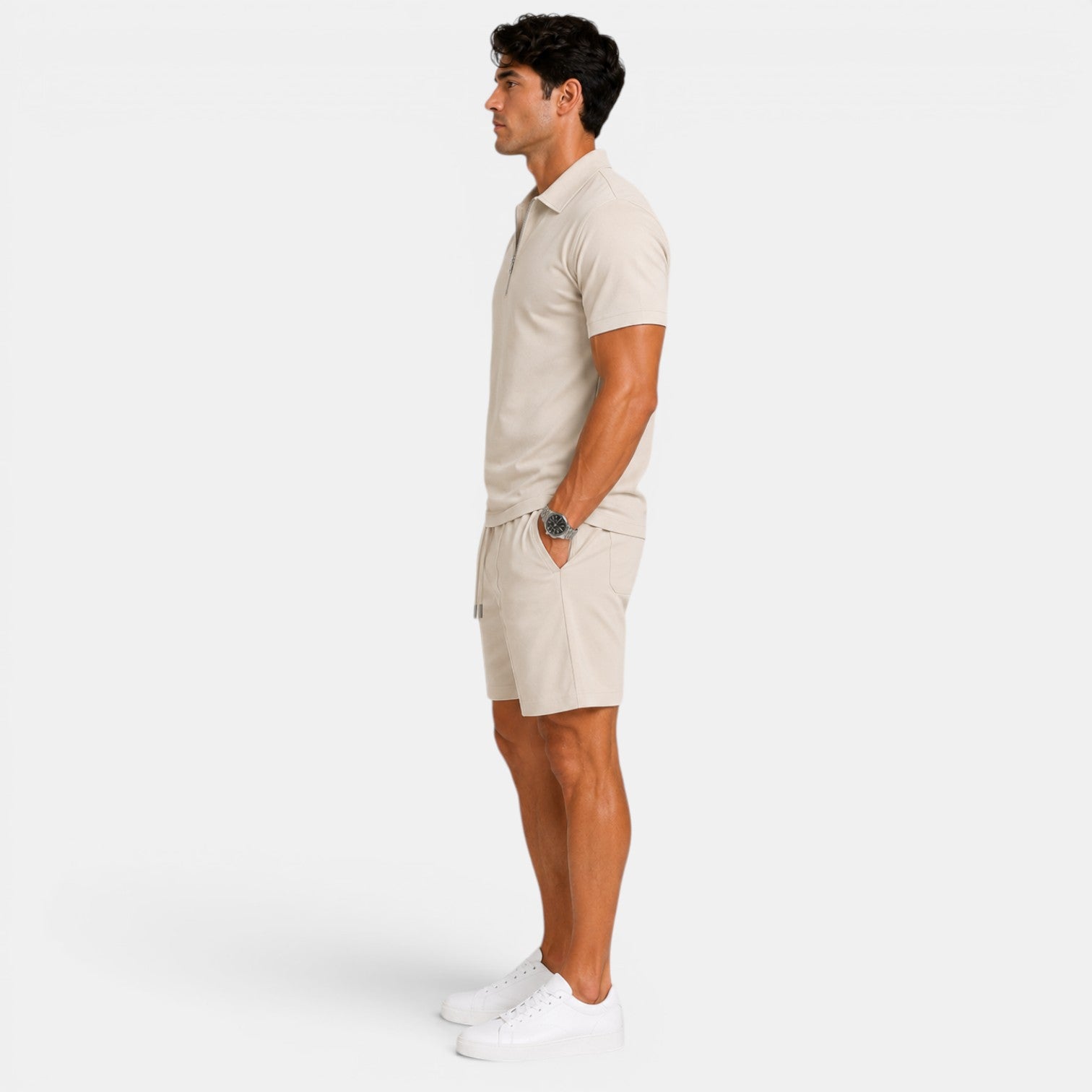 Side view of a man wearing a beige zip-collar polo shirt, matching shorts, and white sneakers against a light gray background.