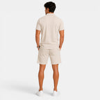 Back view of a man wearing a beige polo shirt, matching shorts, and white sneakers against a light gray background.