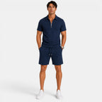 Man wearing a navy blue zip-collar polo shirt with matching drawstring shorts and white sneakers, standing against a light gray background.