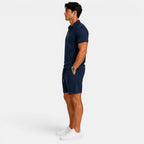 Side view of a man wearing a navy blue zip-collar polo shirt, matching shorts, and white sneakers against a light gray background.