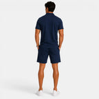 Back view of a man wearing a navy blue polo shirt, matching shorts, and white sneakers against a light gray background.