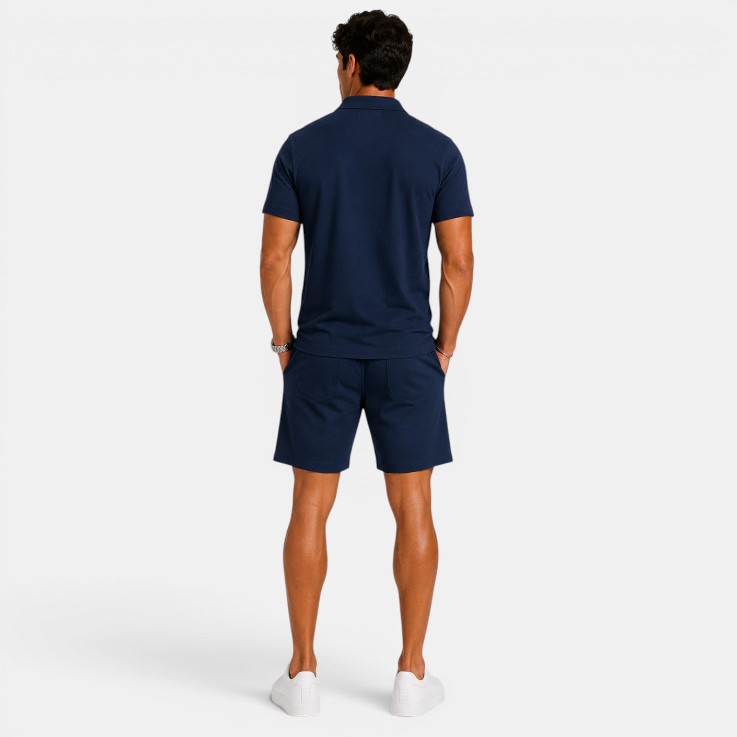 Back view of a man wearing a navy blue polo shirt, matching shorts, and white sneakers against a light gray background.