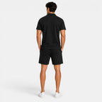 Back view of a man wearing a black polo shirt, matching shorts, and white sneakers against a light gray background.