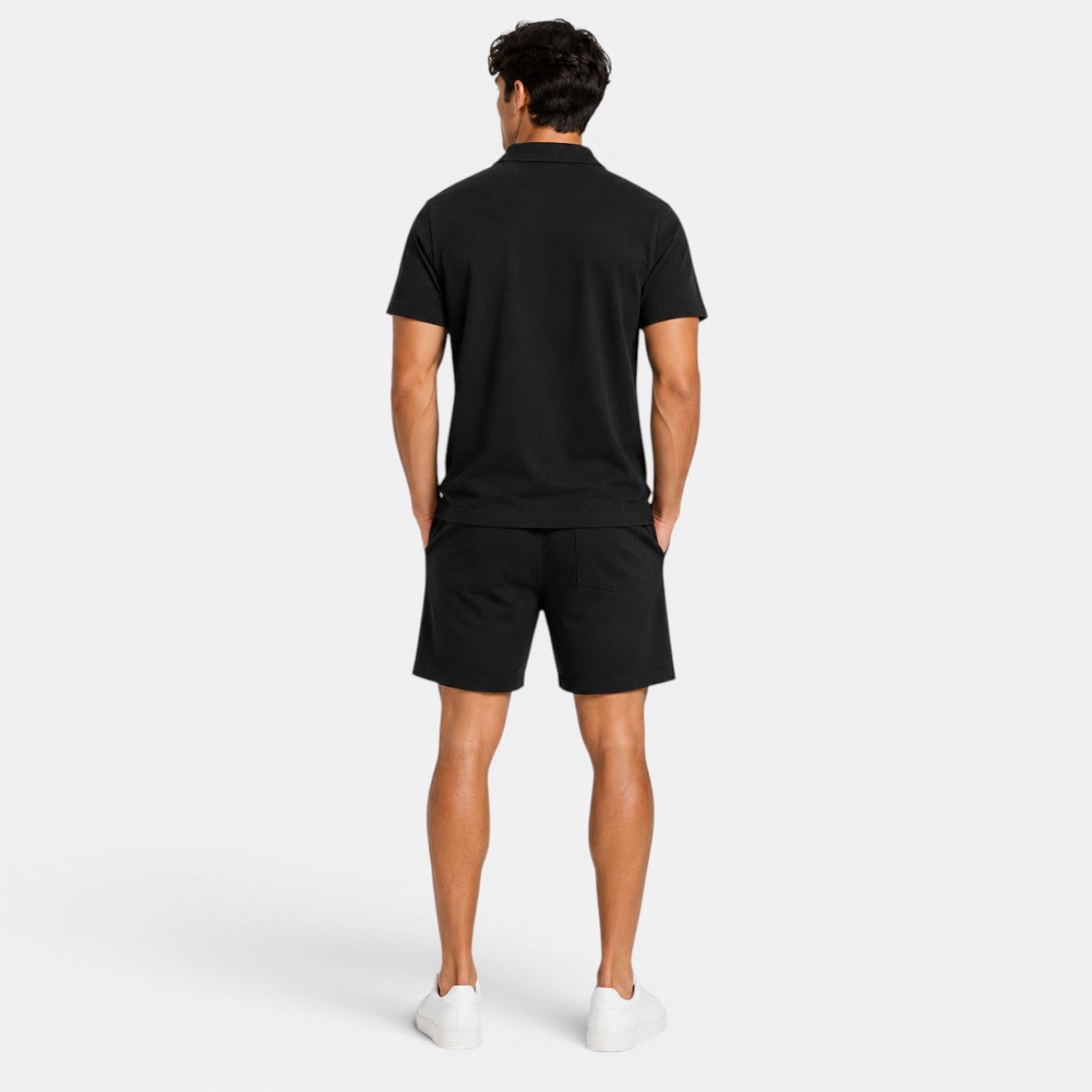 Back view of a man wearing a black polo shirt, matching shorts, and white sneakers against a light gray background.