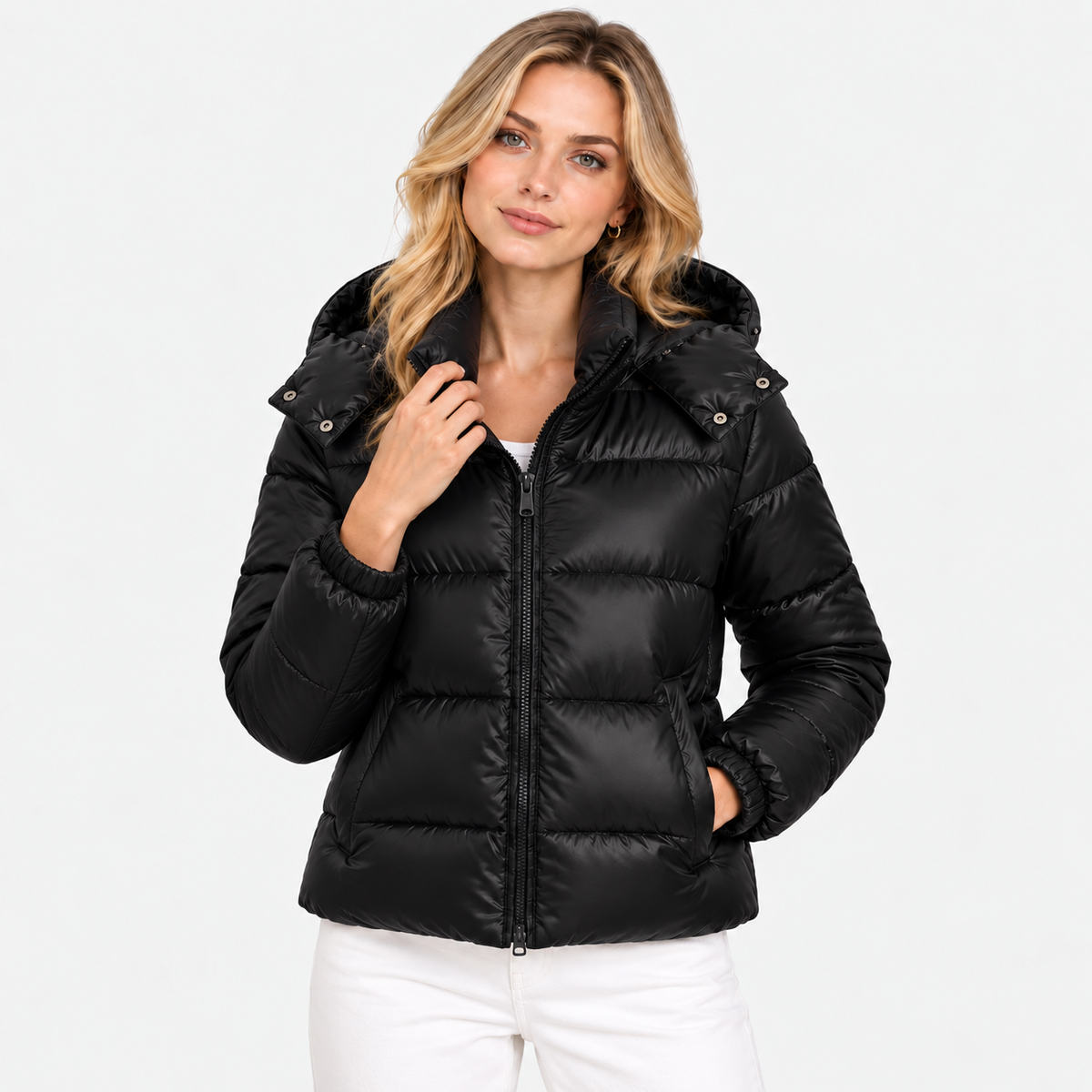 A woman wearing a black hooded puffer jacket with a front zipper and horizontal quilting, paired with white pants, photographed against a clean light gray studio background.