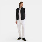 Man wearing a black vest over a white shirt with white pants on a light gray background