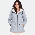 A woman wearing a light gray hooded parka jacket with a cream sherpa-lined hood, black zipper and drawstrings, and front flap pockets, photographed against a plain light gray studio background.