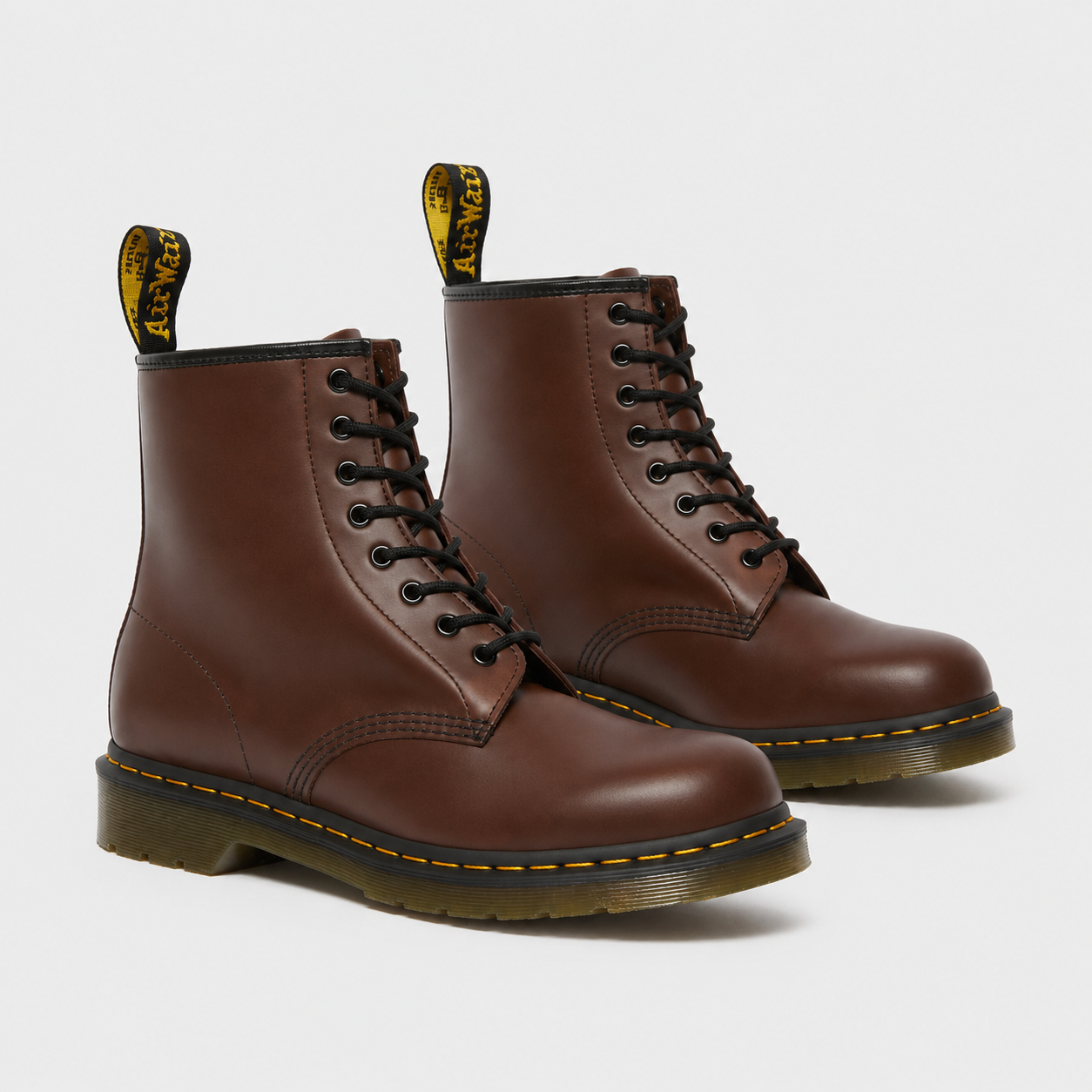 A pair of classic brown leather Dr. Martens 1-8-inch lace-up boots with black laces, black eyelets, and the signature yellow welt stitching and pull tabs, shown on a light gray background.