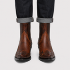 A pair of brown leather western chukka boots with embossed floral detailing on the toes, shown worn with dark cuffed jeans against a light gray background.