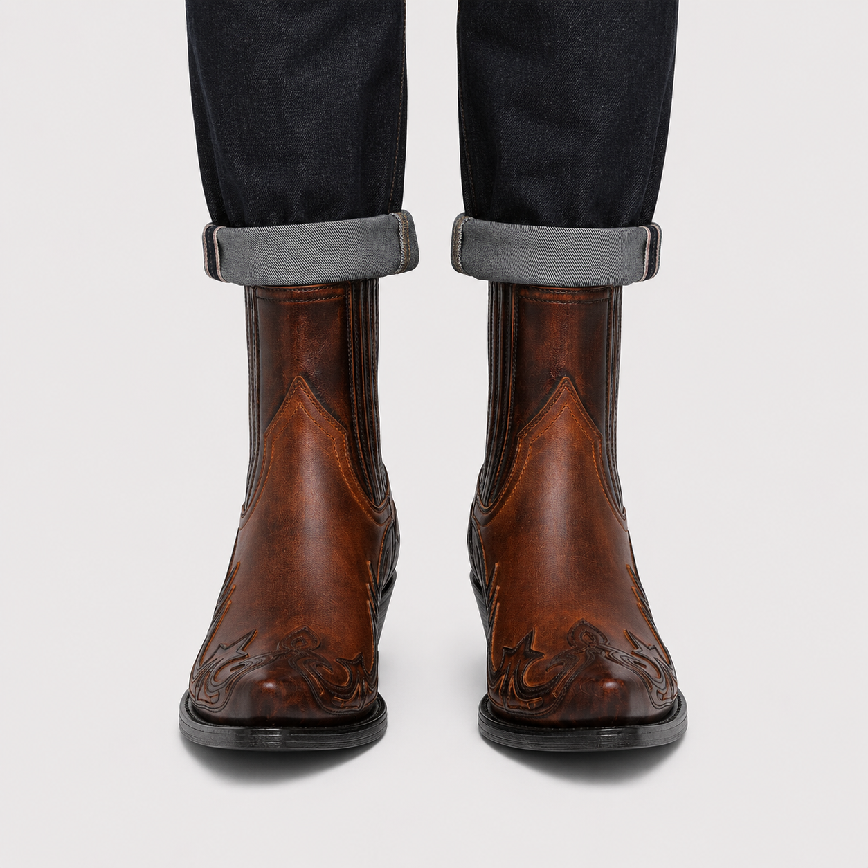 A pair of brown leather western chukka boots with embossed floral detailing on the toes, shown worn with dark cuffed jeans against a light gray background.