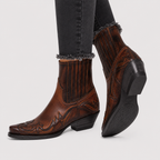A pair of brown western-style leather ankle boots with dark brown and black decorative tooling, worn with dark gray frayed-hem jeans against a light gray background.