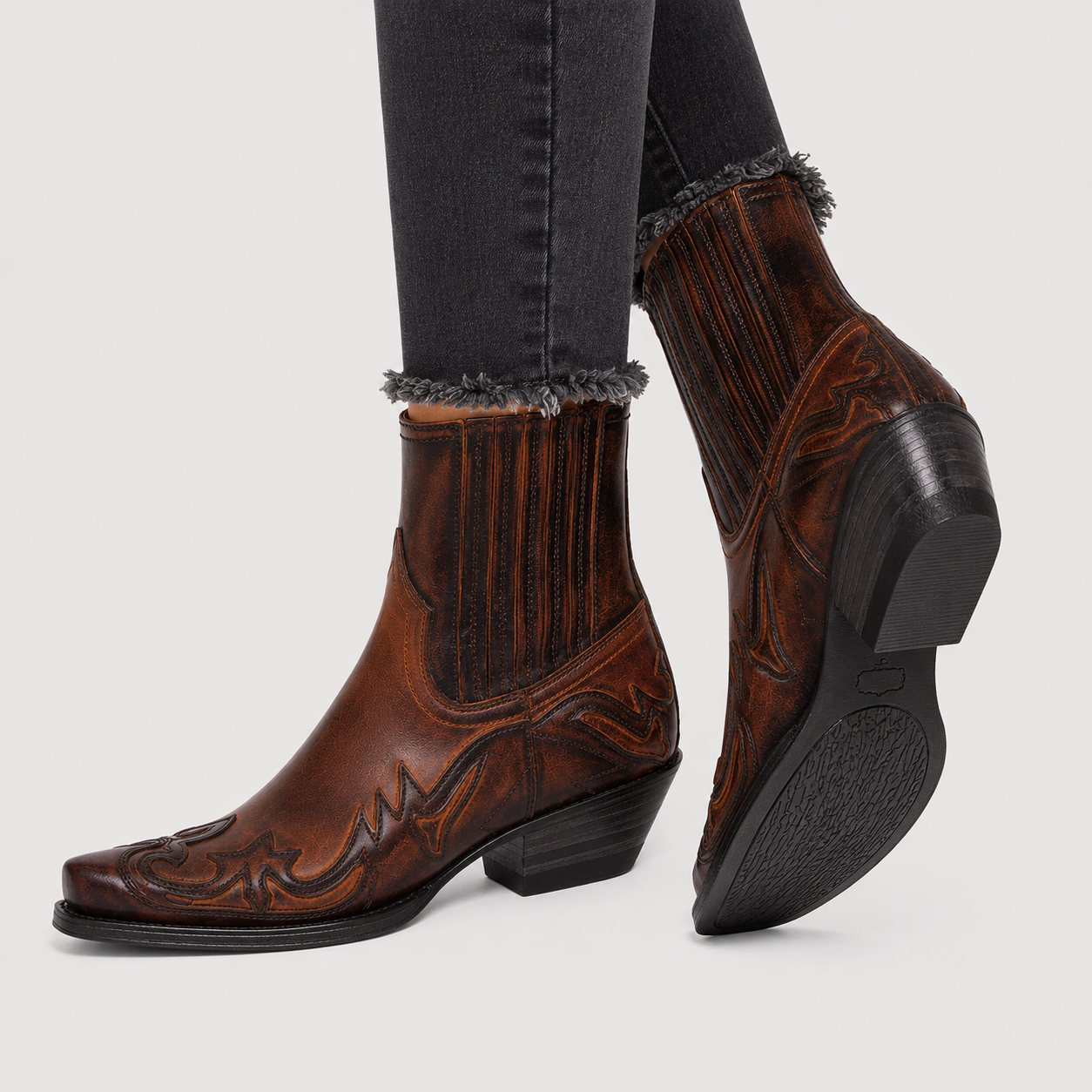 A pair of brown western-style leather ankle boots with dark brown and black decorative tooling, worn with dark gray frayed-hem jeans against a light gray background.