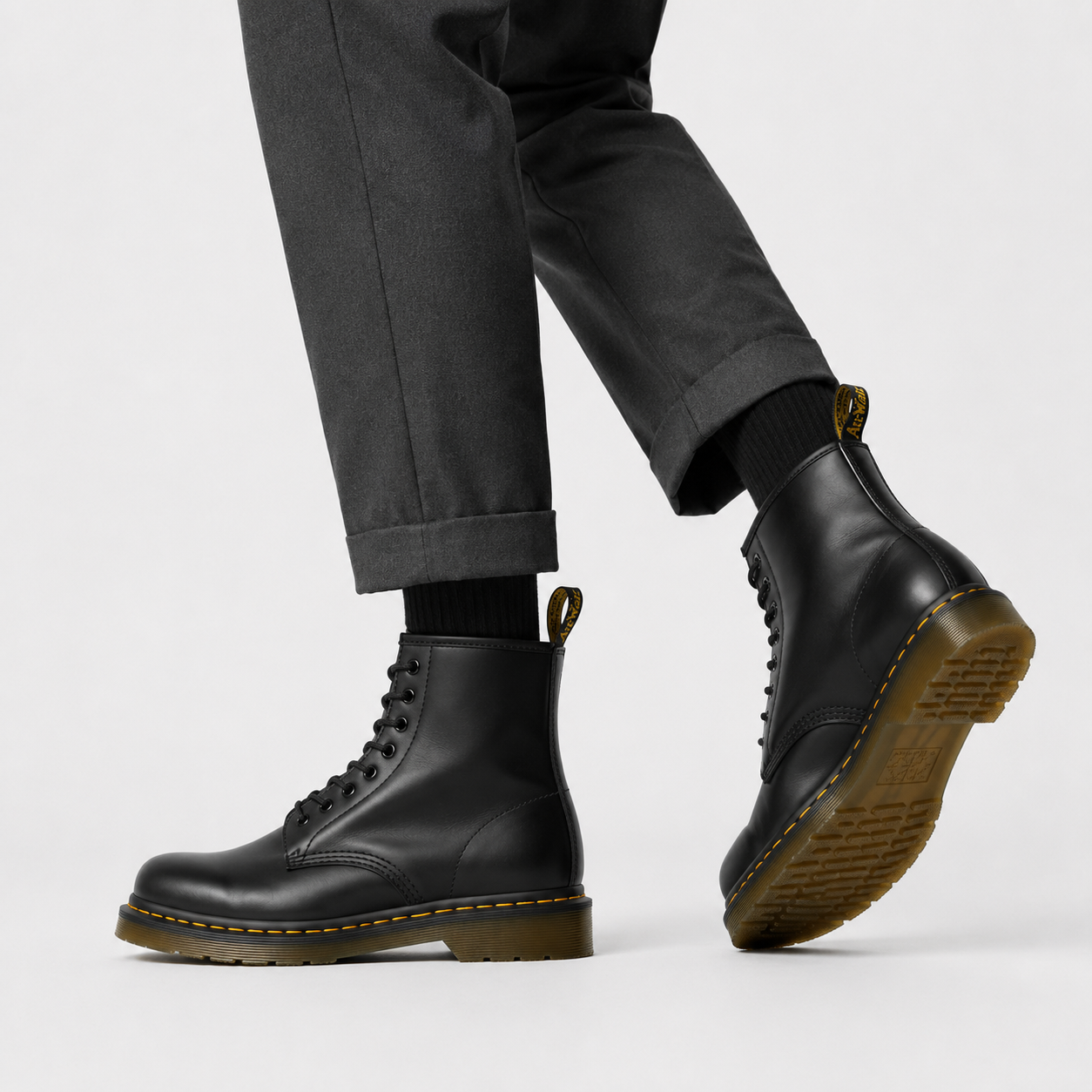 A pair of black leather Dr. Martens 1-8-inch lace-up boots with yellow stitching and gum soles, shown on a model wearing dark gray trousers and black socks against a light gray studio background.