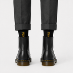 Black leather ankle boots with yellow stitching and brown gum soles, worn with black socks and dark gray trousers, shown from the back on a light gray background.