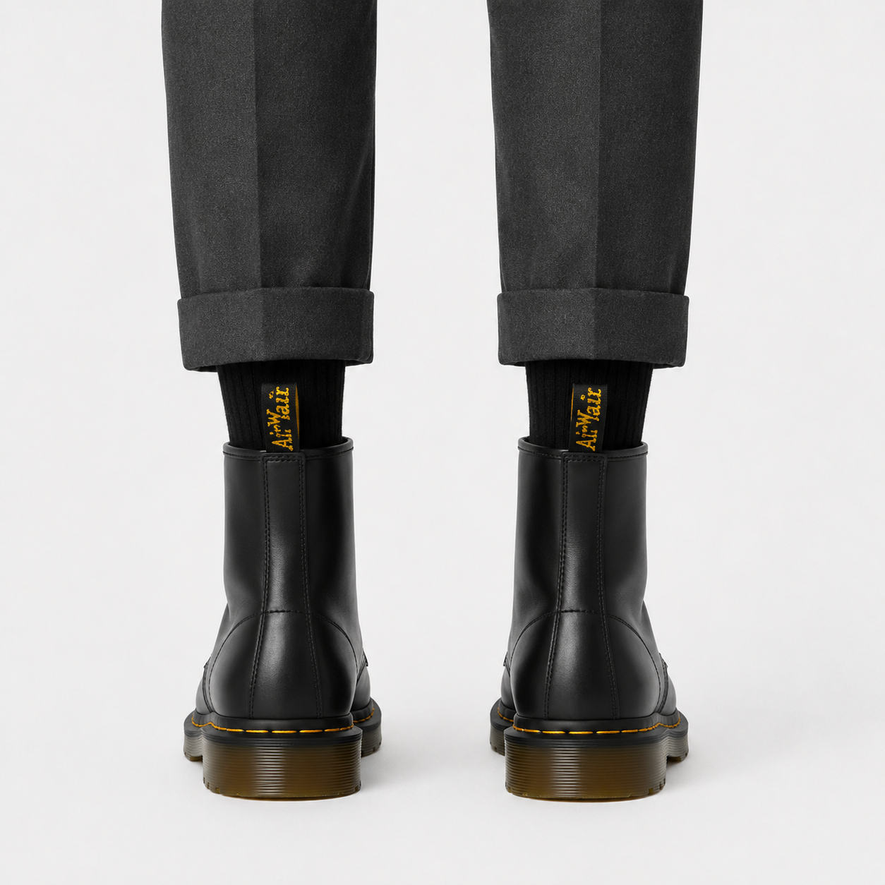 Black leather ankle boots with yellow stitching and brown gum soles, worn with black socks and dark gray trousers, shown from the back on a light gray background.