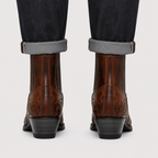 Rear view of a person wearing brown western ankle boots with dark cuffed jeans, shown against a light gray studio background.
