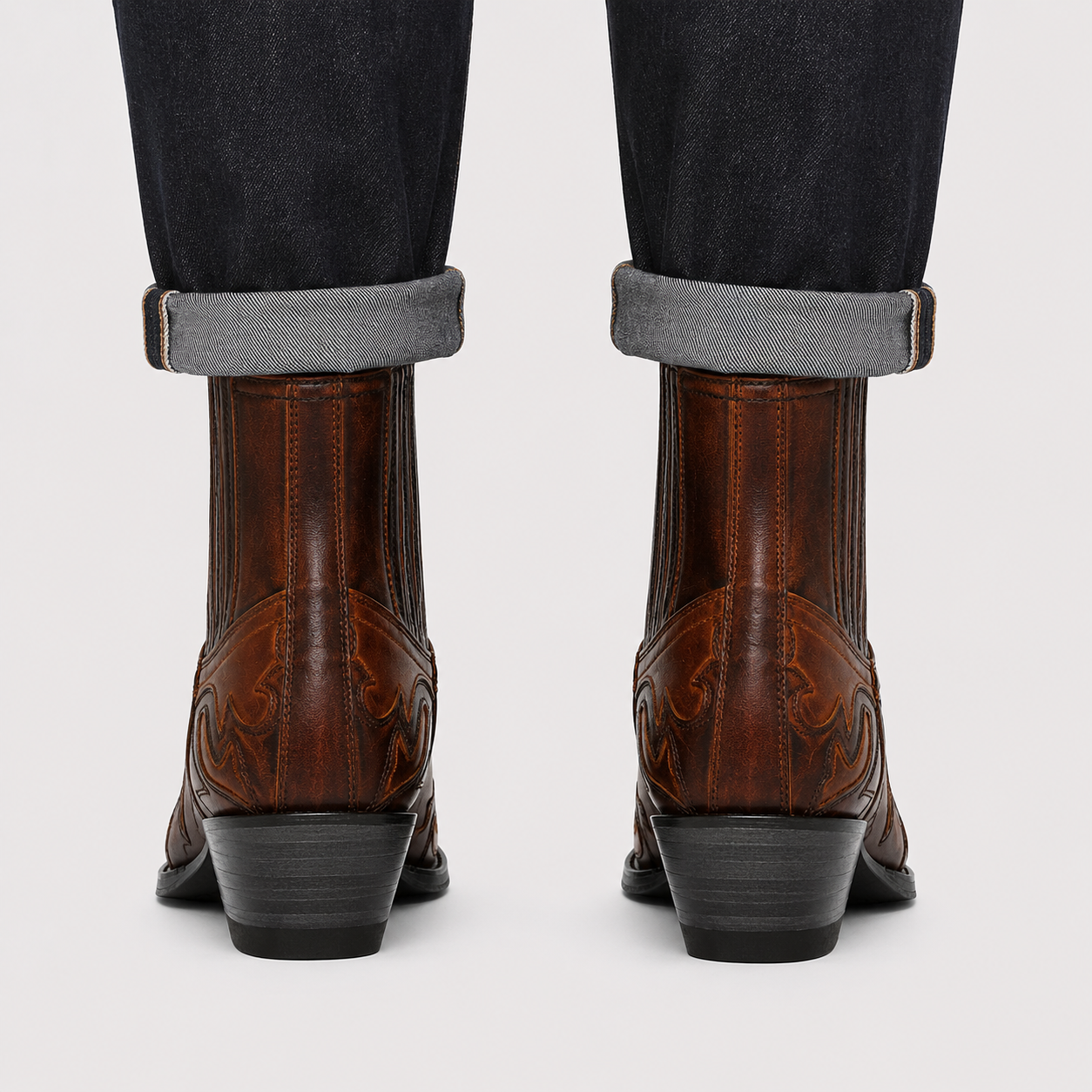 Rear view of a person wearing brown western ankle boots with dark cuffed jeans, shown against a light gray studio background.
