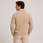 A man shown from the back wearing a beige cable-knit sweater and matching beige trousers against a plain light gray studio background.