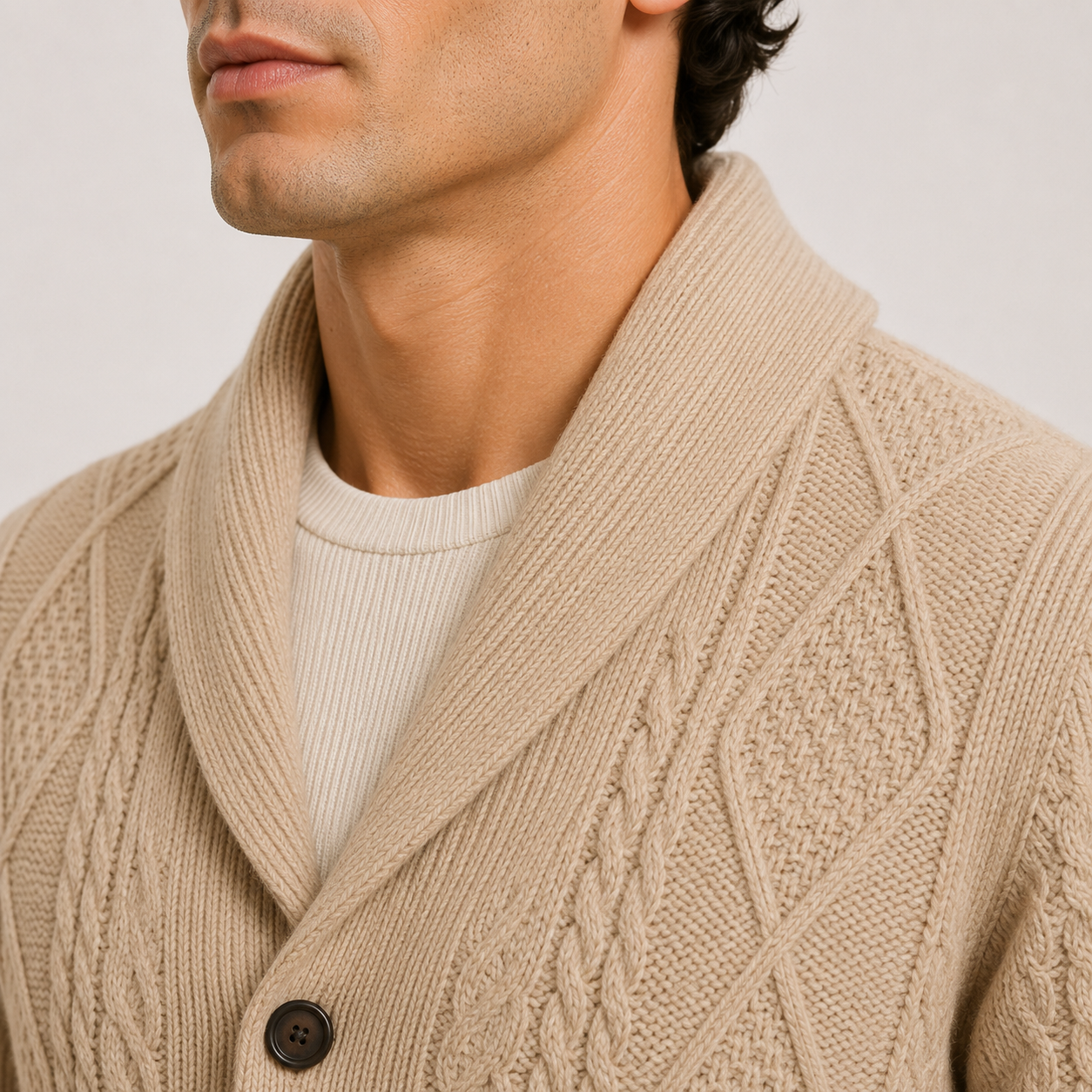 Close-up of a man wearing a beige cable-knit cardigan with a wide collar over a cream ribbed shirt, against a light gray background.