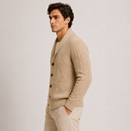 A man wearing a beige cable-knit cardigan over a cream shirt and beige trousers, standing in profile against a light gray studio background.
