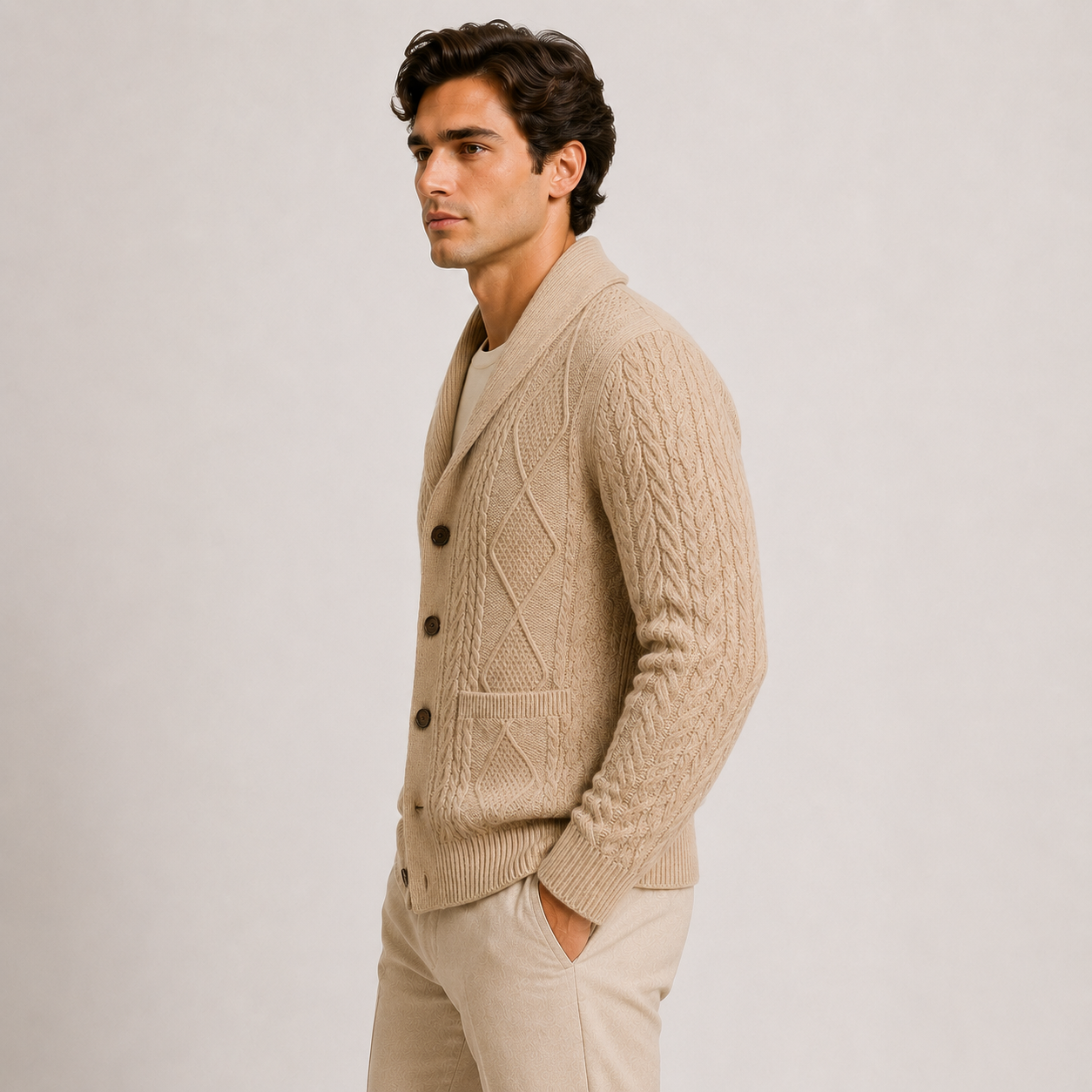 A man wearing a beige cable-knit cardigan over a cream shirt and beige trousers, standing in profile against a light gray studio background.