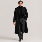 A man modeling a long black double-breasted overcoat over a black turtleneck sweater, dark gray trousers, and black dress shoes, photographed against a light gray studio background.