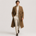 A man modeling a long brown double-breasted overcoat over a cream turtleneck sweater and cream trousers, walking against a light gray studio background.