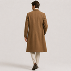 A man viewed from behind wearing a mid-length camel brown overcoat, cream trousers, and dark brown dress shoes against a light gray studio background.
