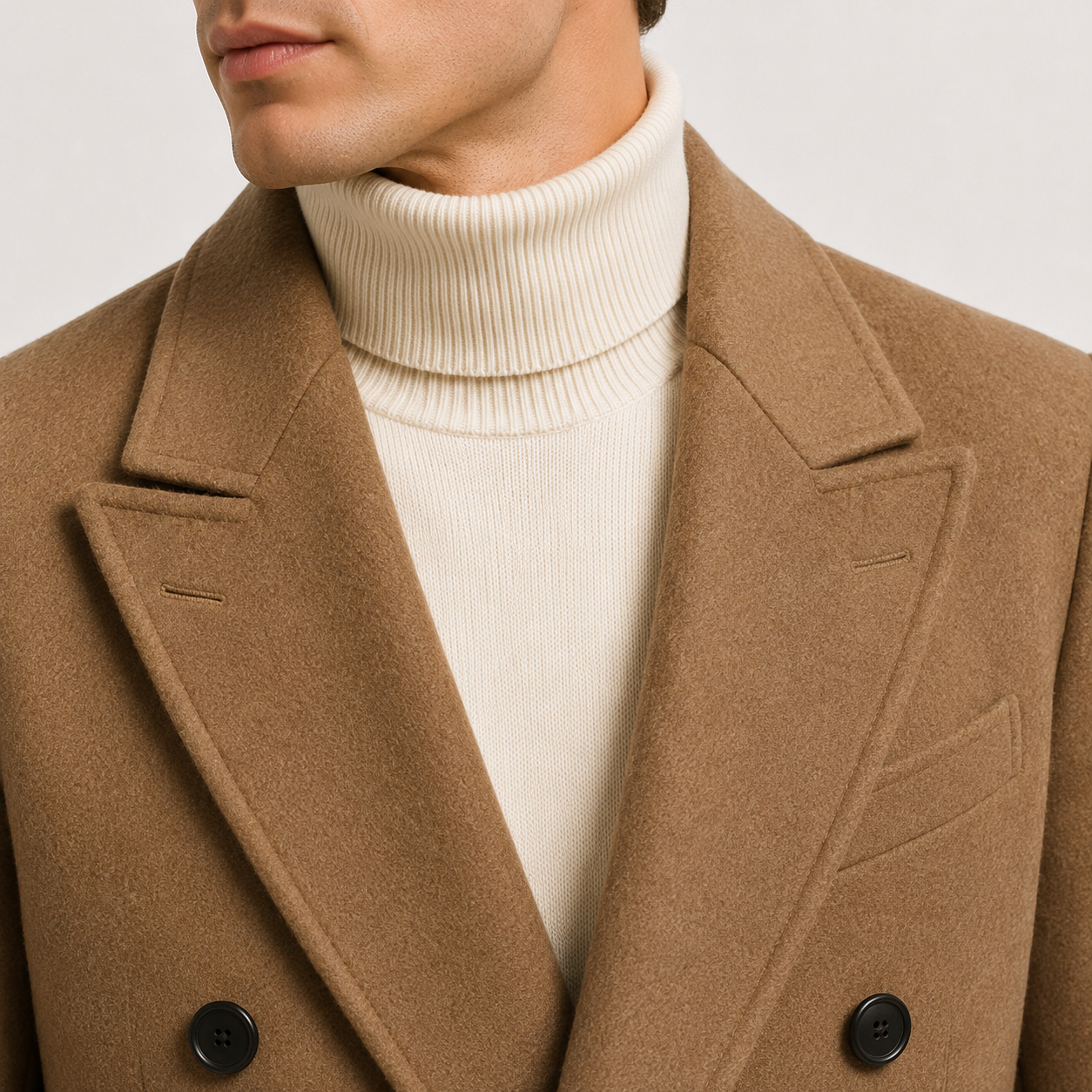 Close-up of a person wearing a camel brown double-breasted coat over a cream ribbed turtleneck sweater, against a light neutral background.