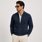 A man wearing a navy blue zip-up suede bomber jacket over a white collared shirt and cream trousers, photographed against a light gray studio background.