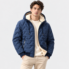 A man wearing a navy blue quilted hooded jacket with a cream sherpa lining, layered over a cream sweater and beige trousers, photographed against a light gray studio background.