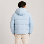 A man viewed from behind wearing a light blue hooded puffer jacket and cream-colored trousers, standing against a neutral light gray studio background.