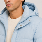 A close-up of a man wearing a light blue hooded puffer jacket over a cream ribbed knit sweater, cropped from the lower face to the chest.