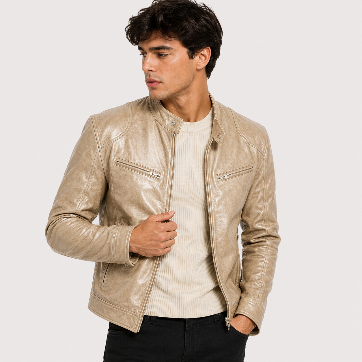 A man wearing a light beige leather moto jacket over a cream ribbed sweater and black pants, photographed against a plain light gray studio background.